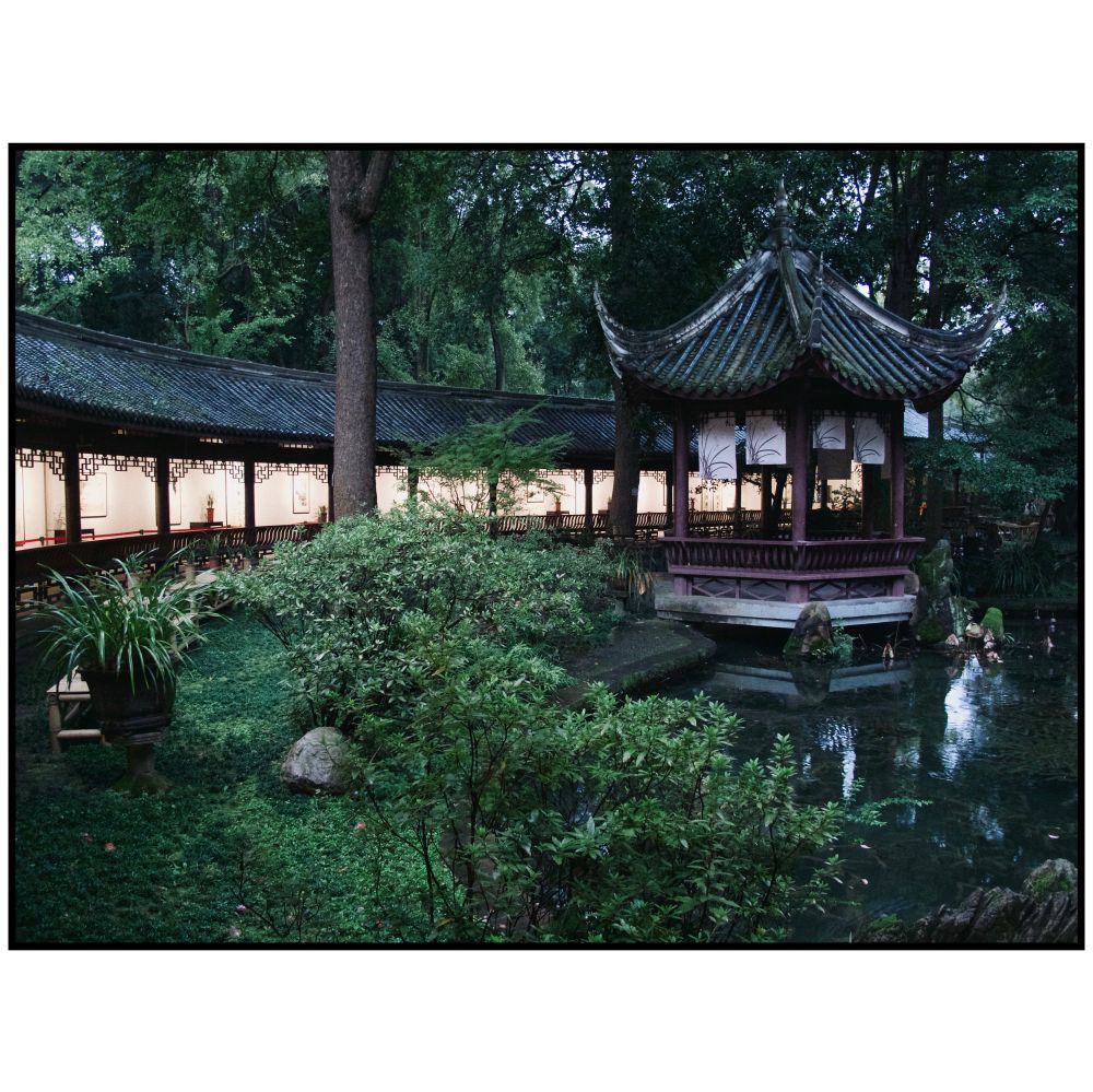 A traditional Chinese pavilion stands beside a reflective pond in Chengdu, illuminated softly from a covered walkway in the background. The scene is calm and contemplative, framed by dense greenery and traditional architecture.