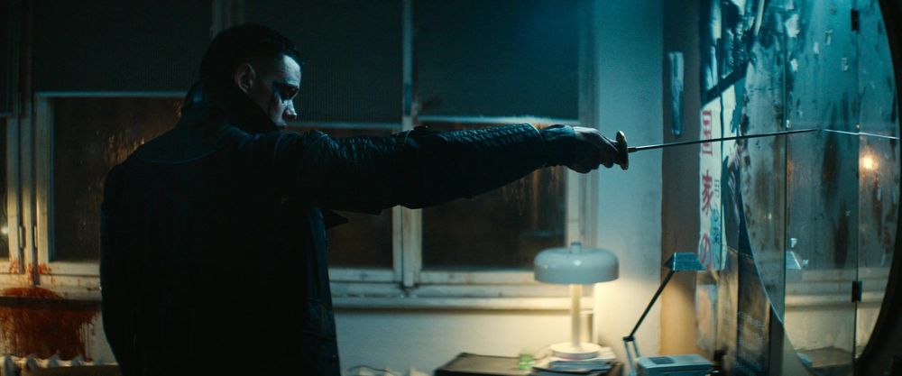 Man in dark leather jacket holding a sword in a dimly lit room with graffitied walls.