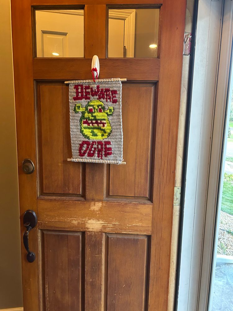 A crochet banner with a picture of Shrek that says “Beware Ogre,” just like in the movie
