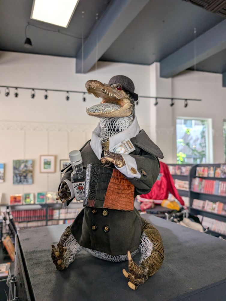 An alligator dressed in a suit and bowler with a bottle of Jim Beam, a briefcase and some ciggys.