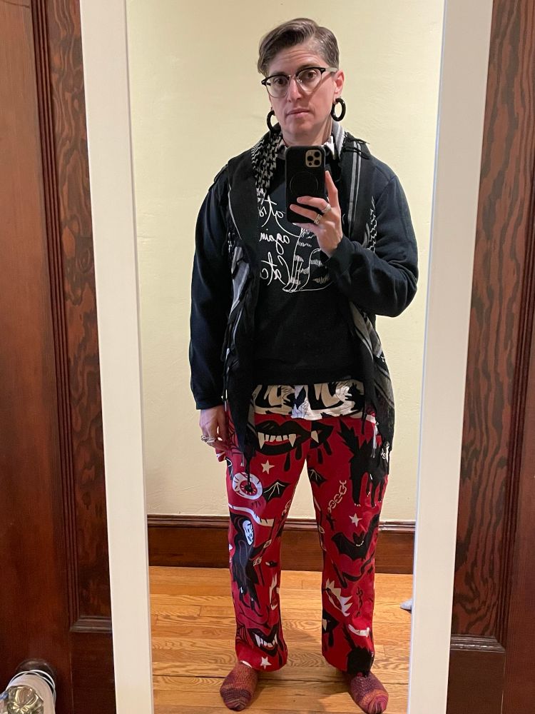 A short haired person stands in front of a full length mirror wearing red, black, and orange handknit socks; bright red painters pants printed with black vampire mouths, bloodshot eyeballs with bat wings, and the grim reaper; a white flannel shirt printed with black outlined hands making shadow puppet animals and the black shadows; a black sweatshirt with a line drawing of an upright sitting cat with script reading “cats against catcalls; a black and white keffiyeh, and large black hoop earrings. Their head is cocked to the side and they are holding their phone in their right hand taking the picture.