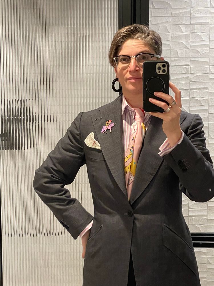 A person stands in a hotel bathroom wearing a hip length, wide lapel grey suit in a modern 1940s style; a pink, purple, and white shirt with French cuffs peeking from the suit sleeves; a pink and yellow silk scarf folded on the bias and tied in a half Windsor knot inside the open shirt collar; a silk pocket square printed like a piece of college ruled paper; and a lapel pin depicting David Bowie and Prince riding a pink unicorn with a purple mane and tail.