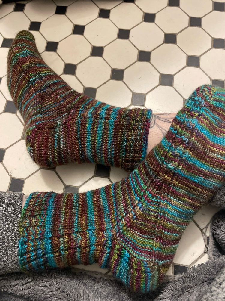 A closeup of two feet in hand knit socks in a variegated brown, green, purple, and turquoise with a cable up the inner leg that splits at the gusset, crossed against a white tiled floor with small black squares at the intersection of the white tiles.