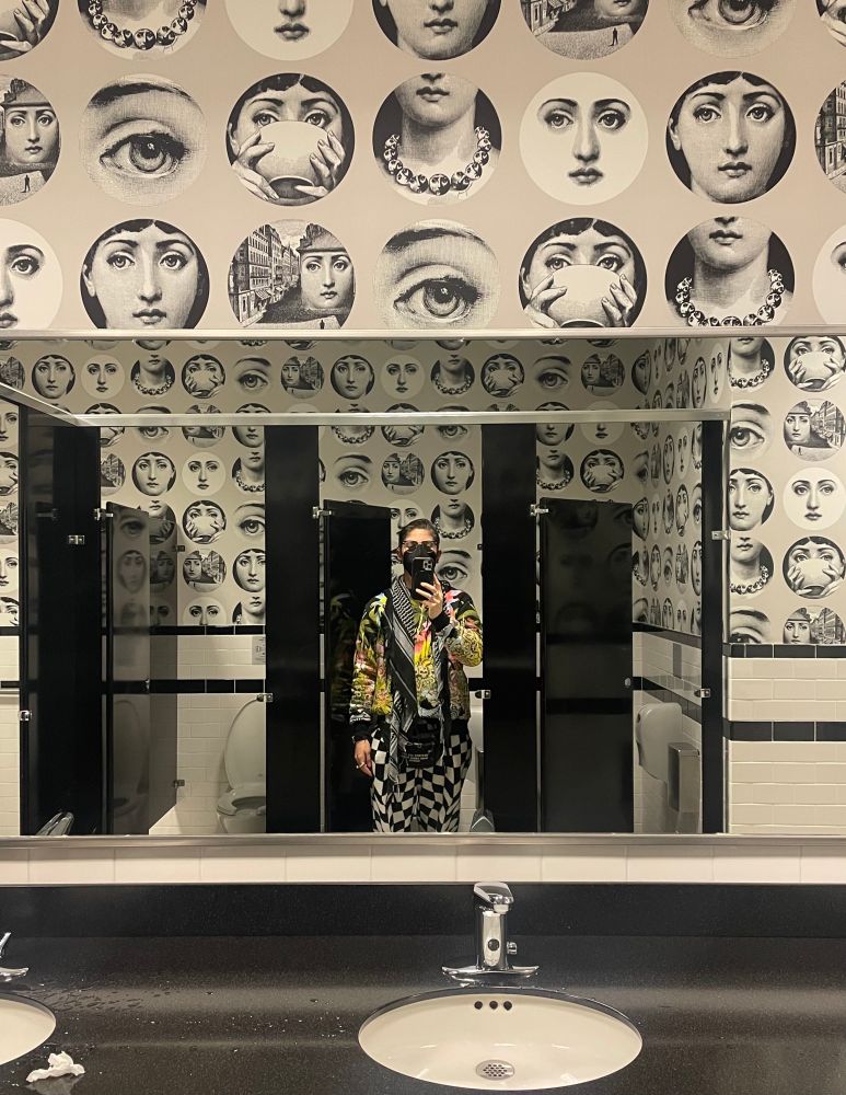 A person stands in an open bathroom stall with an open stall on either side taking a bathroom selfie. A black and ecru wallpaper with large circles featuring stylized women’s faces including a closeup of an eye with an arched brow; a nose to collar neck wearing a moon phase necklace; eyes with arched brows above two hands sipping from a teacup; and feminine features on a white circle. The person is wearing a boldly colored sweatshirt printed with parrots, carnivorous plants, and skulls; black and white checked sweatpants; and a black and white keffiyeh around their neck.