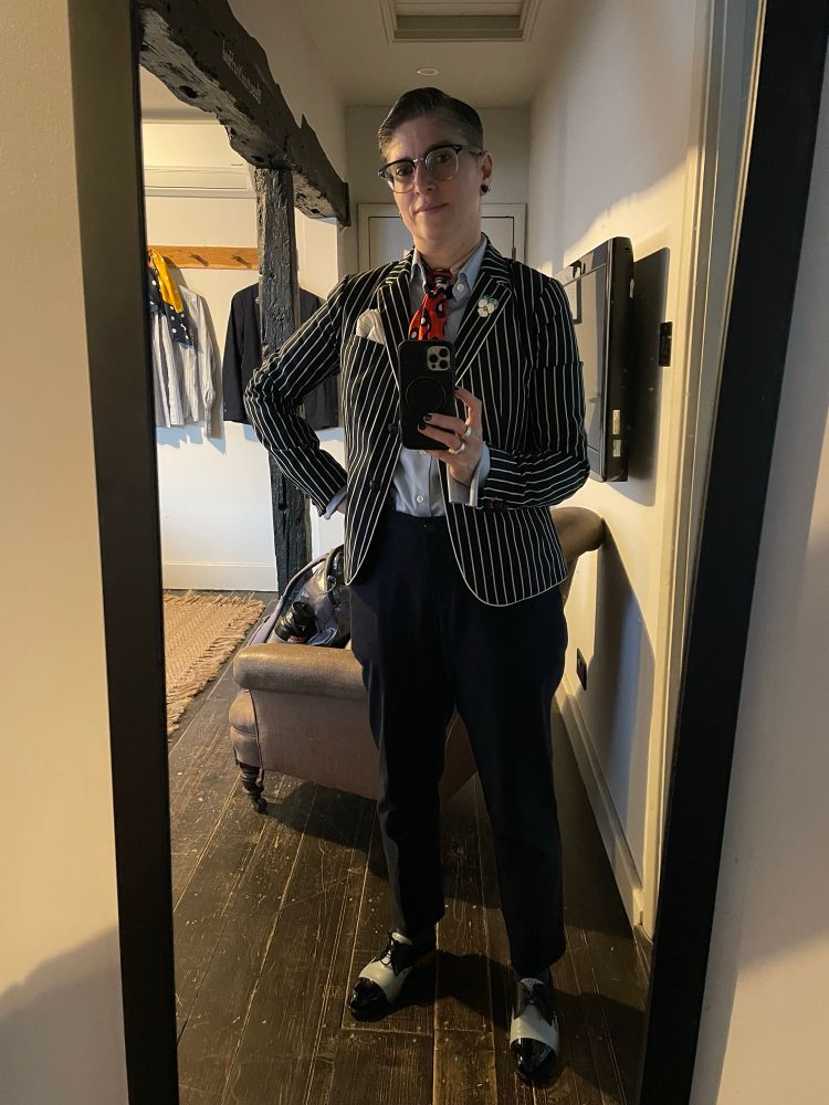 A nonbinary person stands before a full length hotel mirror wearing a navy and white striped schoolboy blazer, a light blue French cuff dress shirt, a red, navy, and cream polka dot neckerchief, navy speckled trousers, and navy and light blue dress shoes.