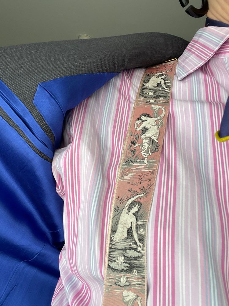 A close up of the open jacket revealing a bright blue lining, the shirt stripes, and pink silk braces featuring topless naiads bathing.
