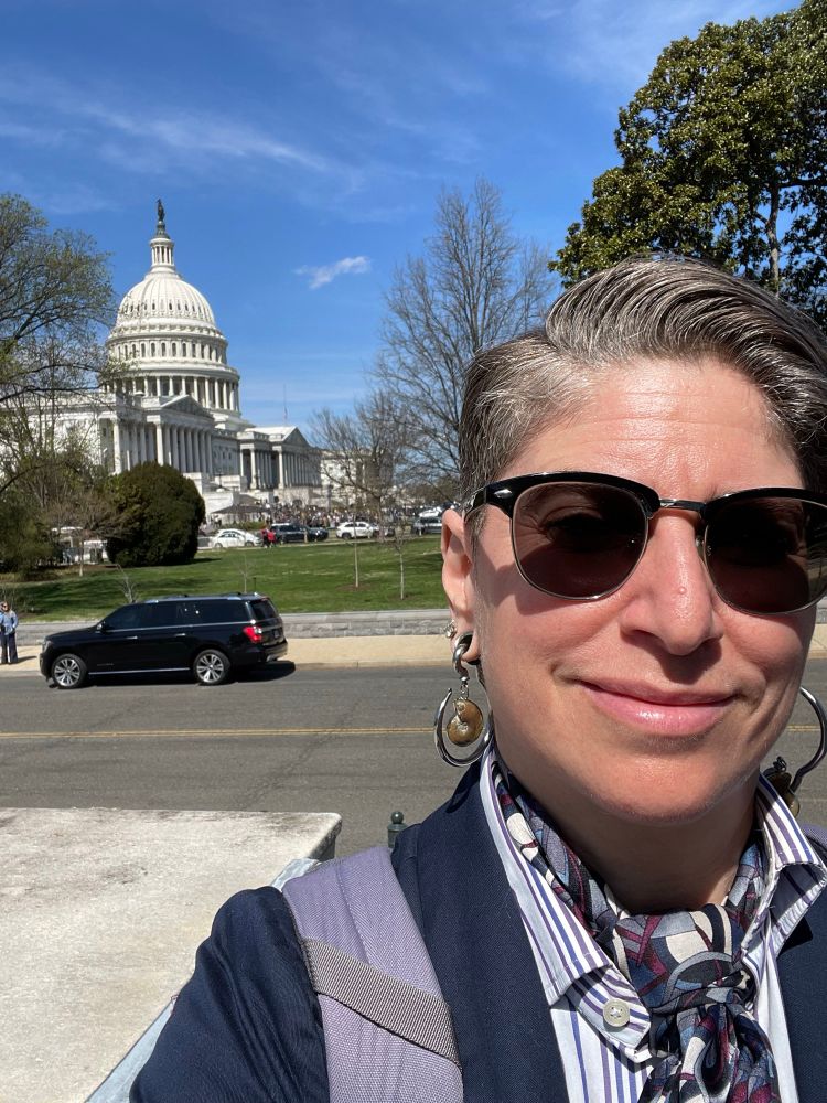 A 47 year old human smiles into the camera in a blue blazer, white dress shirt with blue and purple stripes, a blue and purple patterned silk scarf tied in a full Windsor knot, and sunglasses, with the US Capitol dome in the background over their right shoulder. 