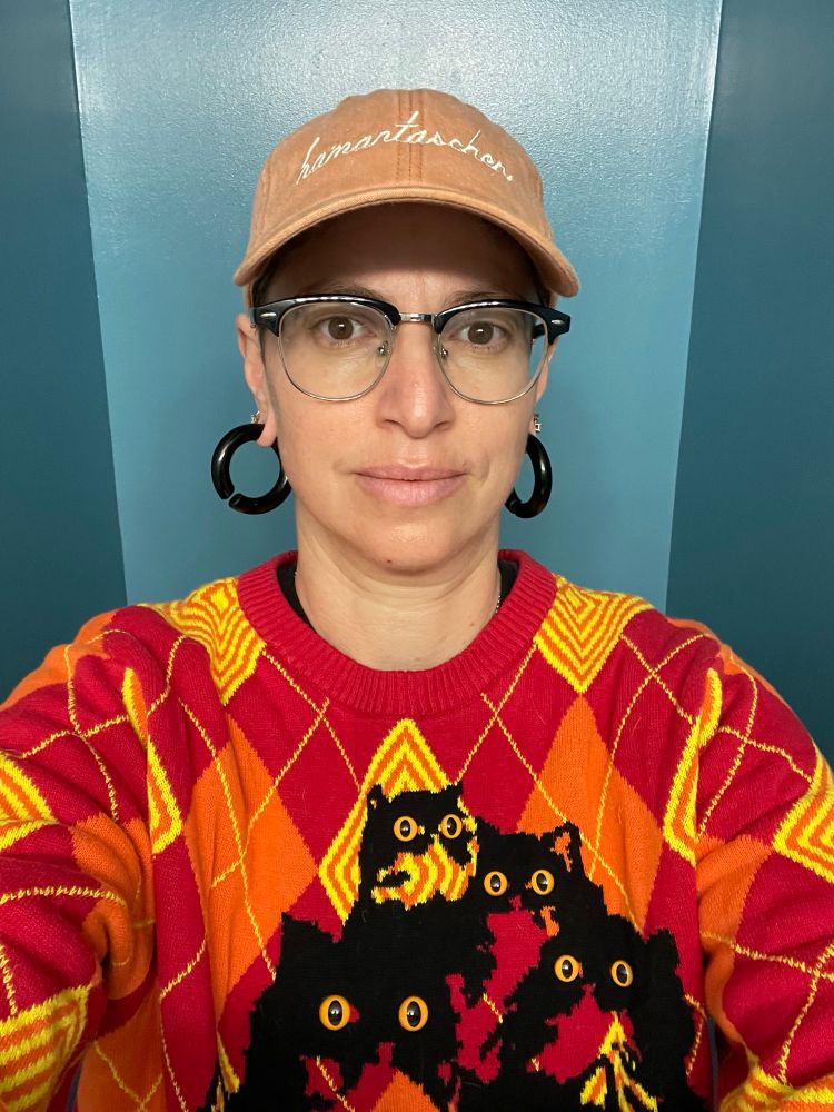 A person stands in a teal alcove wearing a red, orange, and yellow argyle sweater with a pyramid of black cat silhouettes with large orange beaded eyes, black hoop earrings, glasses, and a pale orange hat that says “hamantaschen” (a cookie for the Jewish holiday of Purim) in white script. They have a faint smile and a scratch on their upper lip from their baby nibbling and on their chin from their 4 year old nibbling. Too much roughhousing!