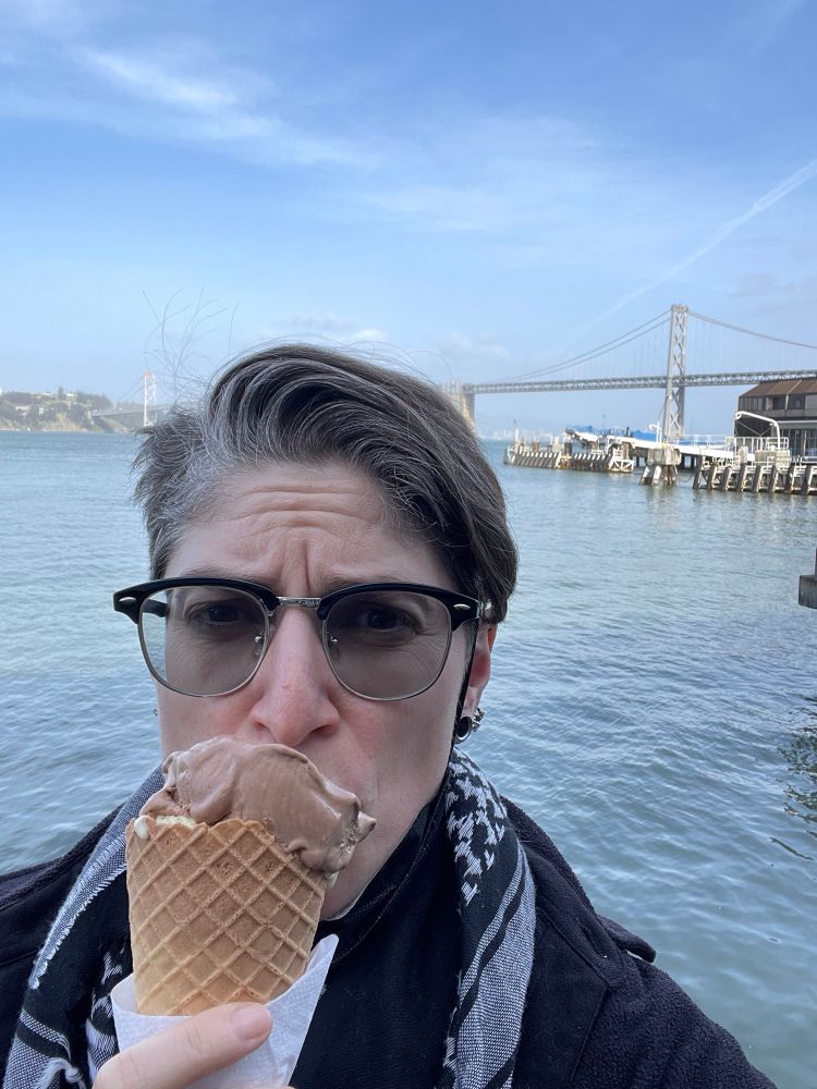 A selfie of a person with a wrinkled forehead, black hoodie, and keffiyeh eats a large waffle cone of dark chocolate and smoked sea salt ice cream (there’s a hidden scoop of peanut butter curry below) in front of water with the Bay Bridge in the background.