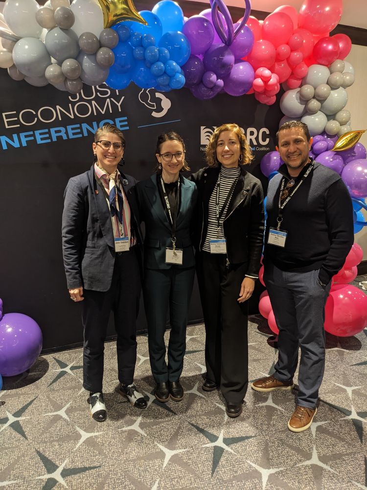 Same person stands with 3 other members of the Local Return Community Investment Cooperative team in front of a balloon arch with the words “Just Economy Conference” in the background.
