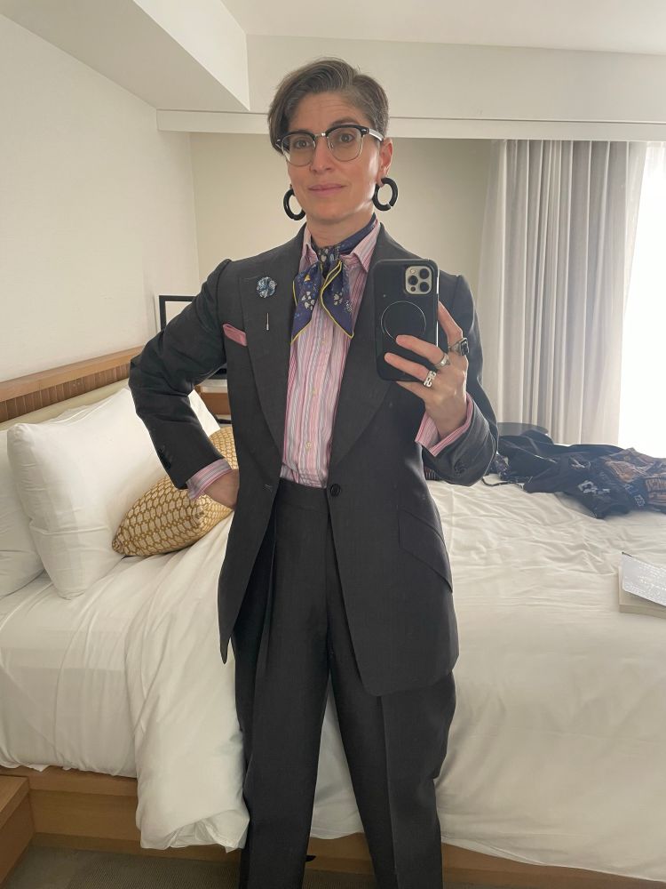A person stands before a mirror wearing a grey two-piece suit with a long jacket and wide lapels; a pink, fuchsia, blue, and white  striped French-cuff dress shirt, a pink pocket square, a blue fabric flower stick lapel pin, a blue floral silk scarf tied on the bias in a square knot inside the open shirt collar, large black hoop earrings, with earlobe length undercut hair.