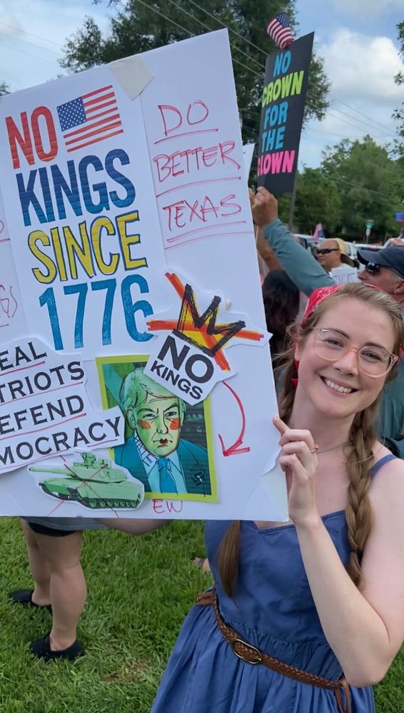 A smiling (beautiful) young woman (Emily, my fiancé) with braided hair and glasses holds a large handmade protest sign reading “NO KINGS SINCE 1776” with colorful, bold letters and an American flag graphic. The sign features a satirical cartoon of Donald Trump with clown-like makeup and a yellow crown crossed out with the words “NO KINGS” taped over it. Other elements include phrases like “REAL PATRIOTS DEFEND DEMOCRACY” and “DO BETTER TEXAS,” along with a red arrow pointing to the cartoon and the handwritten word “EW.” Behind her, more protestors and American flags are visible.