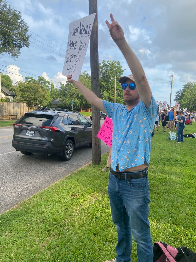 Me standing at a roadside protest (No Kings Day), holding up a white sign in one hand that reads “WHAT WOULD MAKE YOU QUESTION HIM?” in bold black letters and raising my other hand in a peace sign. I’m wearing blue sunglasses, a beige cap, jeans, and a light blue shirt decorated with small yellow flowers. Behind me, protestors line the sidewalk holding various signs, and a car drives by on the road.