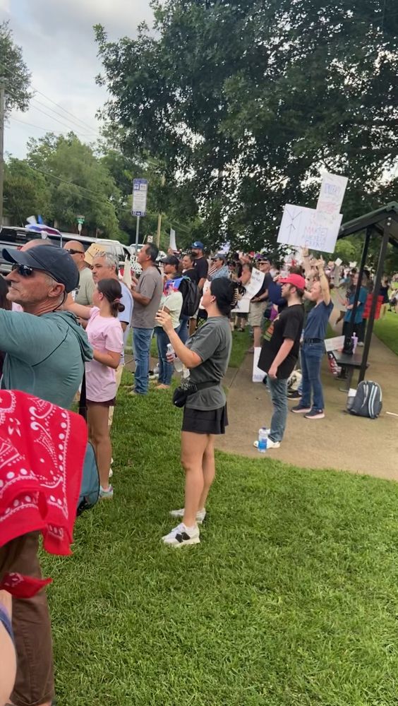 A wide shot of a large, peaceful protest crowd standing on a grassy sidewalk. People of various ages and backgrounds face the street, many holding up handmade signs. One reads “MAKE AMERICA GREAT” with “AGAIN” crossed out. Others are partially obscured. A woman in a gray shirt and black skirt stands in the foreground with her phone raised to record. A red bandana is visible in the lower left corner, and several people wear hats, sunglasses, or patriotic clothing. A tree canopy and a small bus stop shelter are visible in the background.