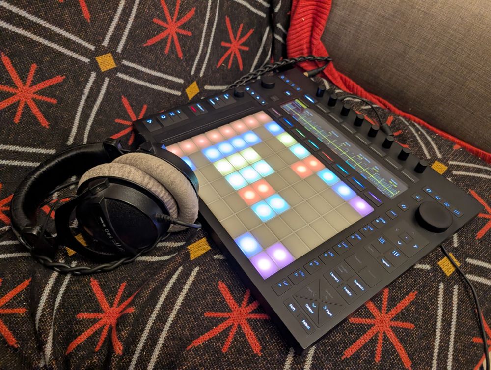 An Ableton Push 3 music box thing with a project on it, the pads and screen are lit up in different colours, there is a pair of Beyerdynamic DT770 headphones next to it, both on a black white red and yellow patterned blanket.