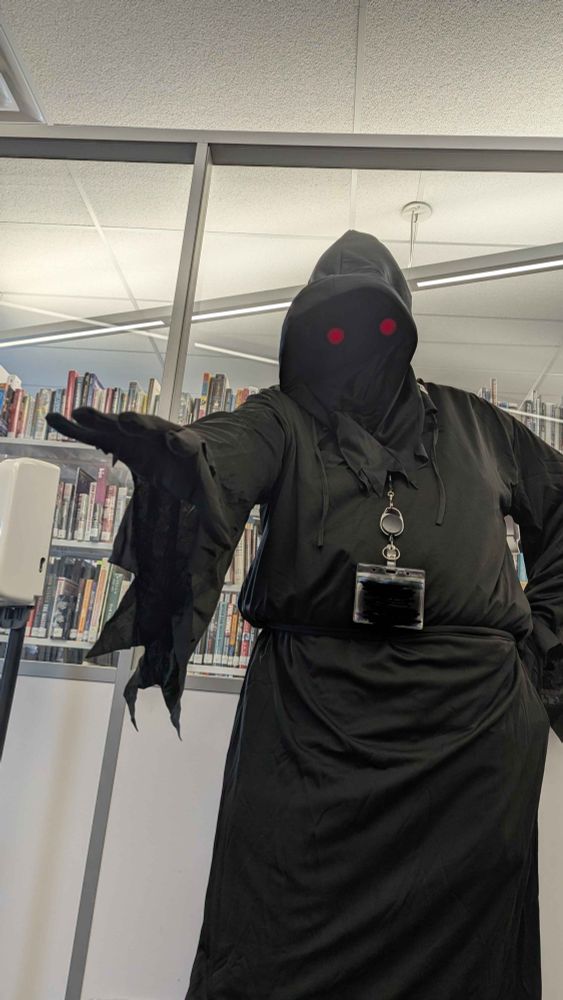 me, a librarian, at my workplace.  i am wearing a grim reaper outfit with glowing red eyes in a dark mask