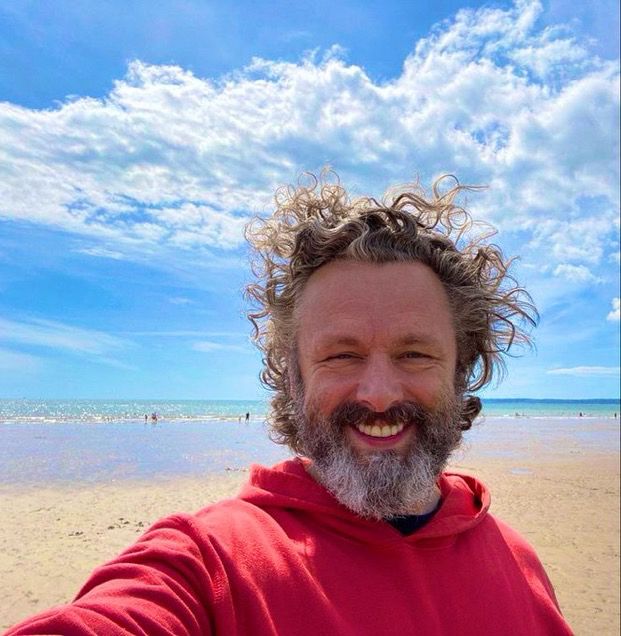 Michael Sheen in his red hoody on a beach.