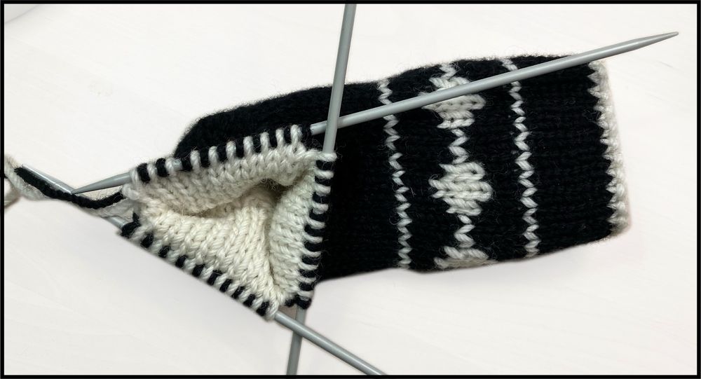 A sock being double knit, which is a black outside layer and a white inside layer, with the stitches arranged alternately on three double-pointed needles. The sock has diamond designs on it in white on the visible black layer, and (because the sock is double knit) the implication is that there are corresponding black designs on the inside white layer.