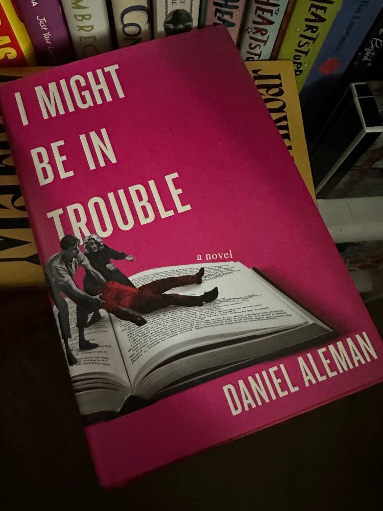 The cover of the book “I Might Be in Trouble” by Daniel Aleman. It had a pink background, and two of the main characters, David and Stacey, are dragging what seems to be a limp body across the page