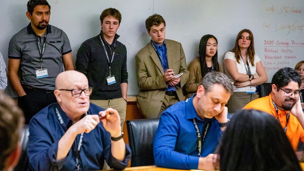 Group of professionals and students in a meeting room listening to a speaker, with a whiteboard in the background filled with handwritten notes.