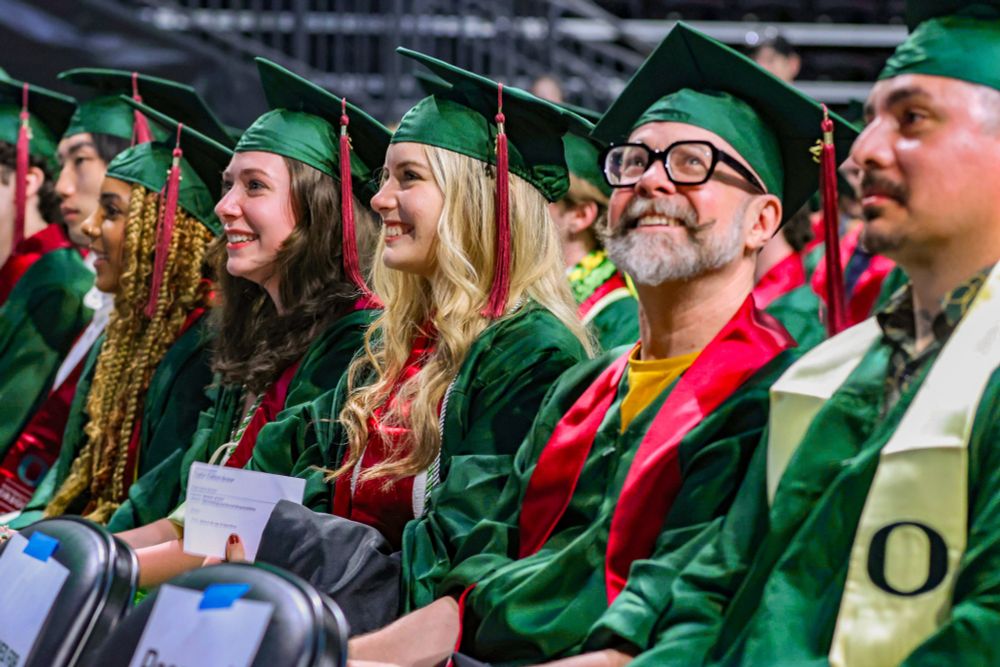 Group of graduates in green robes smiling during a graduation ceremony.

