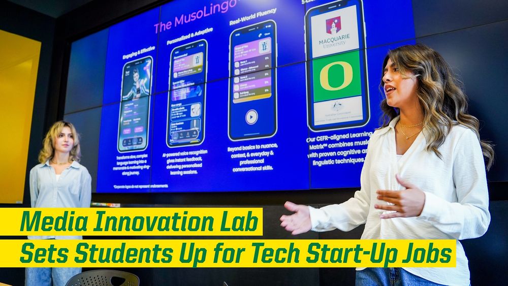 Two individuals presenting an application on a large screen in the Media Innovation Lab. The screen displays app features and information.