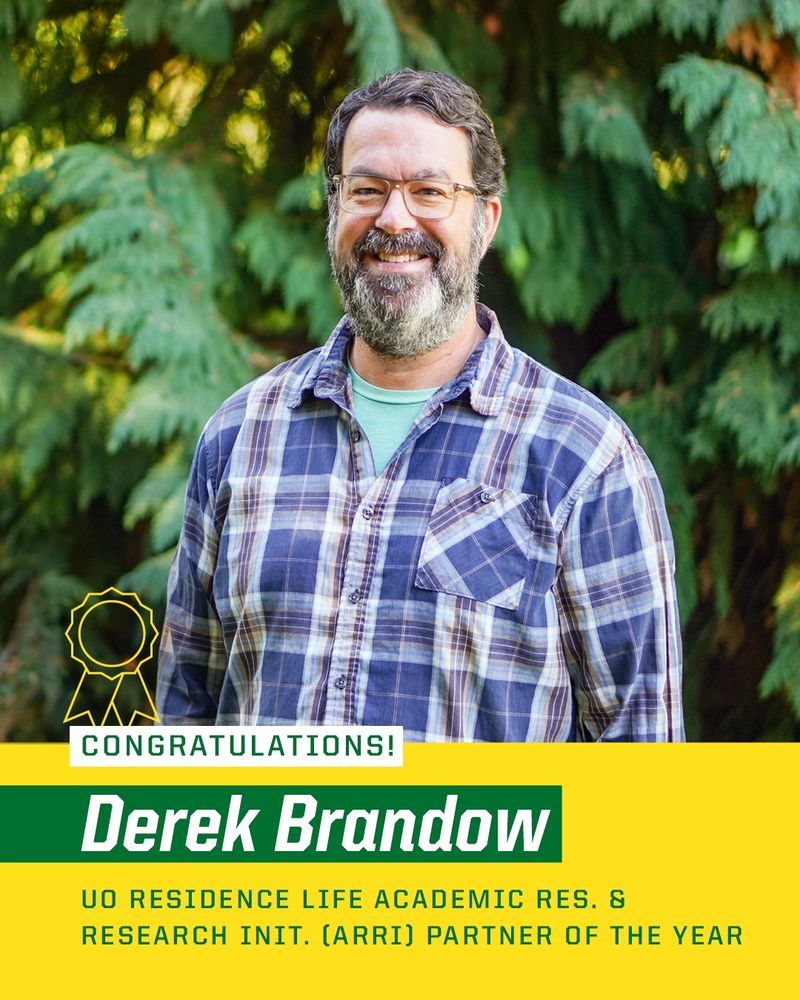 Congratulations Derek Brandow for being awarded the UO Residence Life Academic Res. & Research Init. (ARRI) Partner of the Year, smiling in a plaid shirt with a blurred green foliage background.