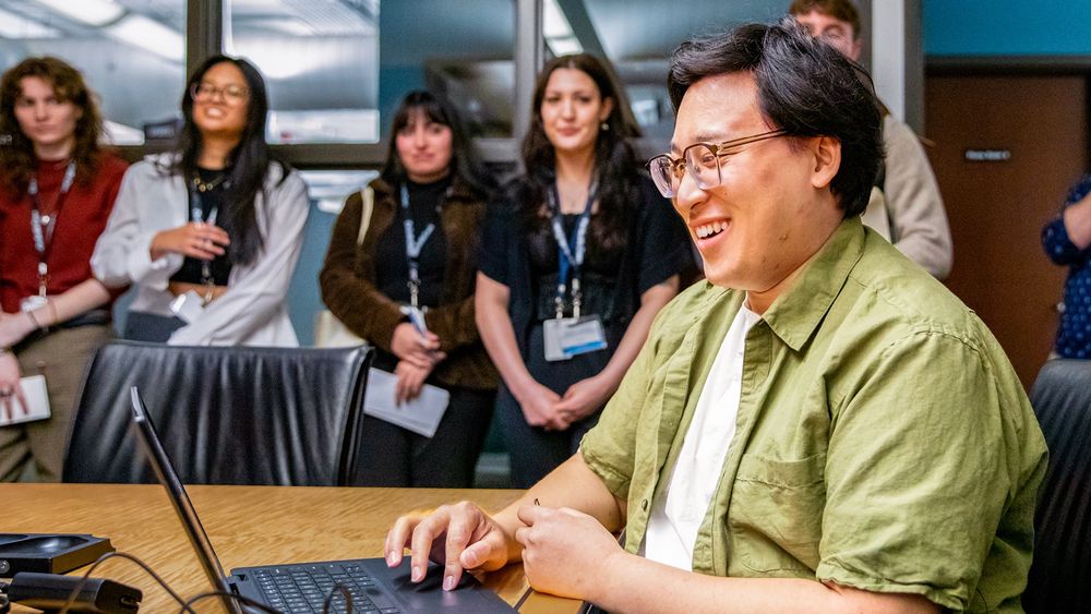 A group of professionals, one seated at a computer laughing joyfully, surrounded by standing colleagues who are smiling and observing in an office environment.