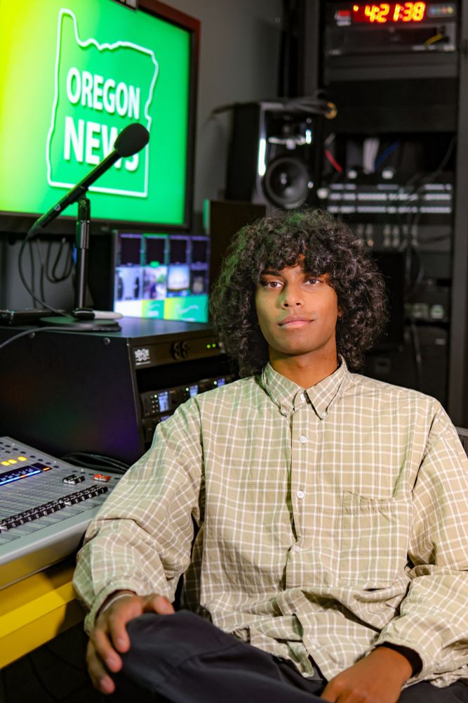 Person sitting in a broadcast studio with "Oregon News" on a screen in the background, surrounded by video production equipment.