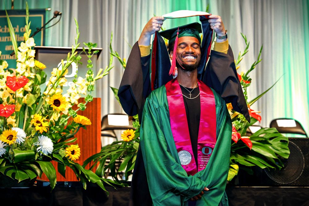 Graduate in regalia on stage at a university commencement ceremony, with a backdrop of vibrant flowers.