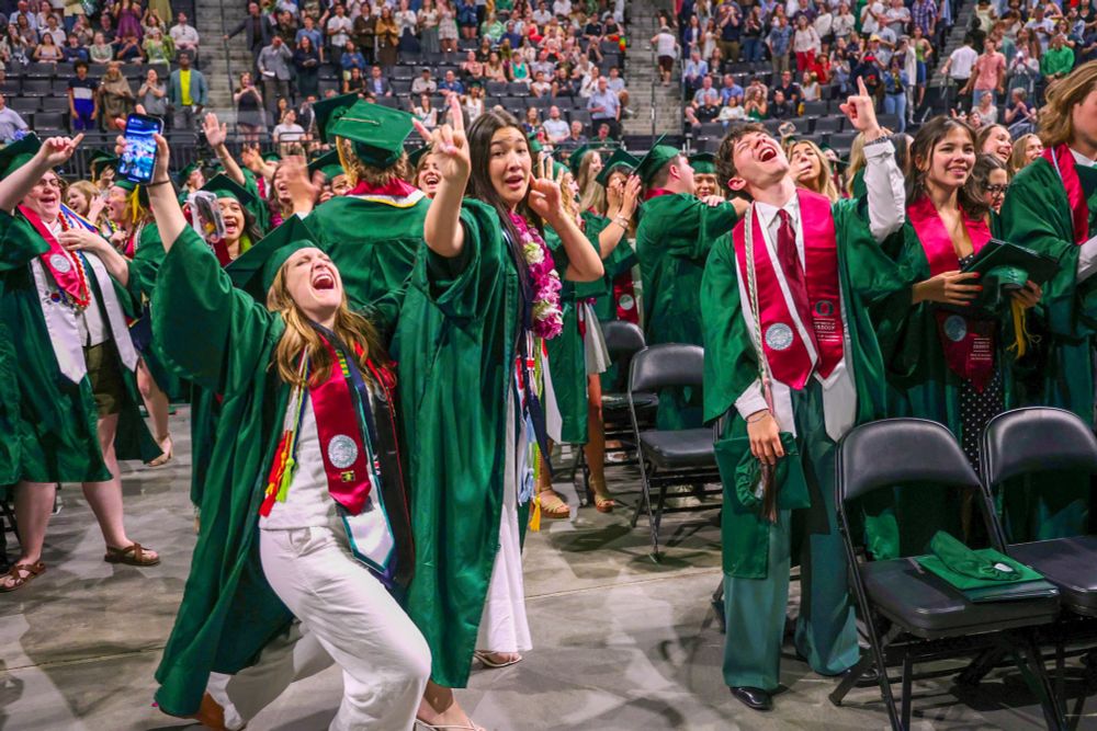 Graduates in green robes excitedly celebrating at their commencement ceremony, some tossing caps in the air, with spectators in the background.


