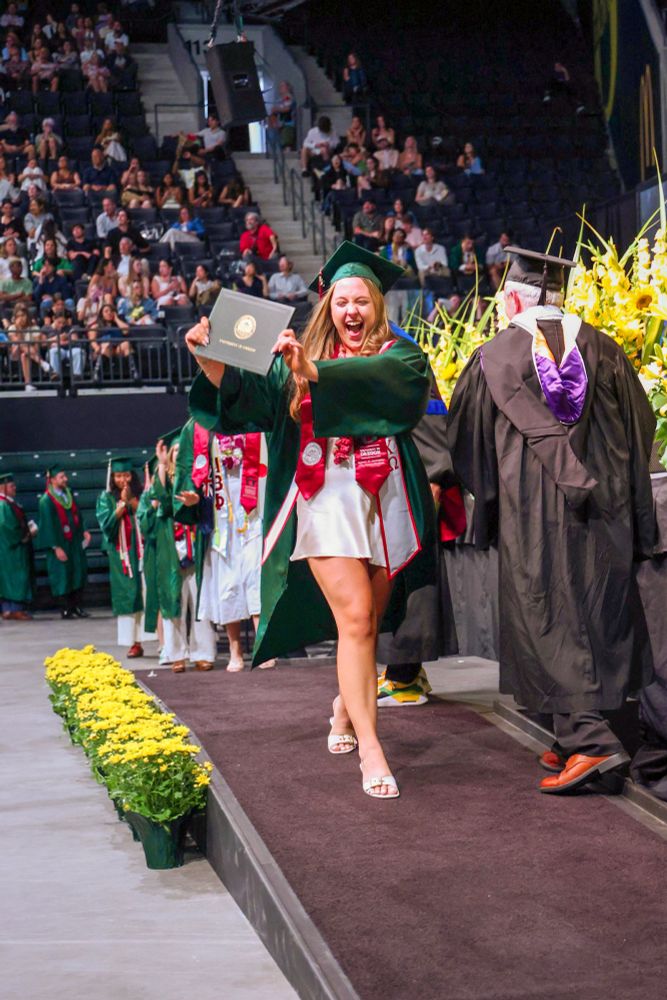 A joyful graduate in a green cap and gown holding a diploma, celebrating while walking across the stage at commencement.