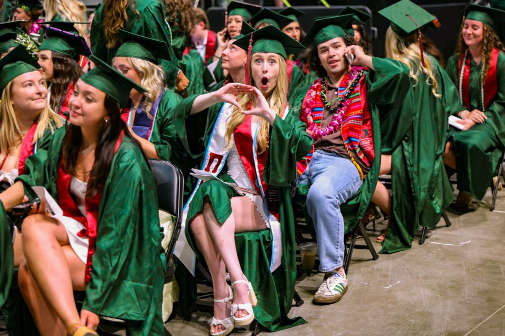 Group of graduates in green robes and mortarboards sitting during a graduation ceremony, looking joyful and excited. Some are wearing decorative sashes and leis.

