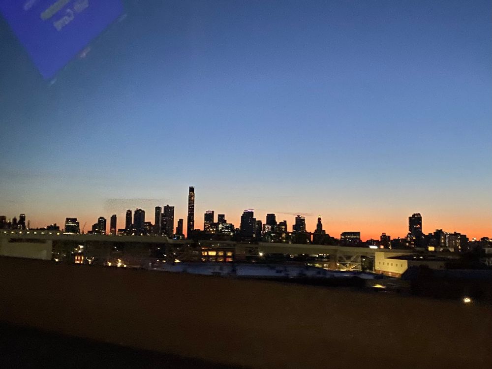 Sun rising over Manhattan from a distance. Sky goes from blue to orange with buildings silhouetted in the foreground. 