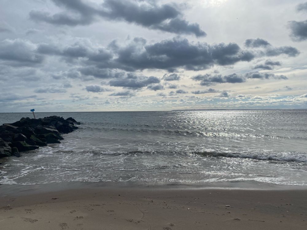 Sun sparkling off the ocean at Brighton Beach, a rock jetty on the left, clouds speckling the sky. 