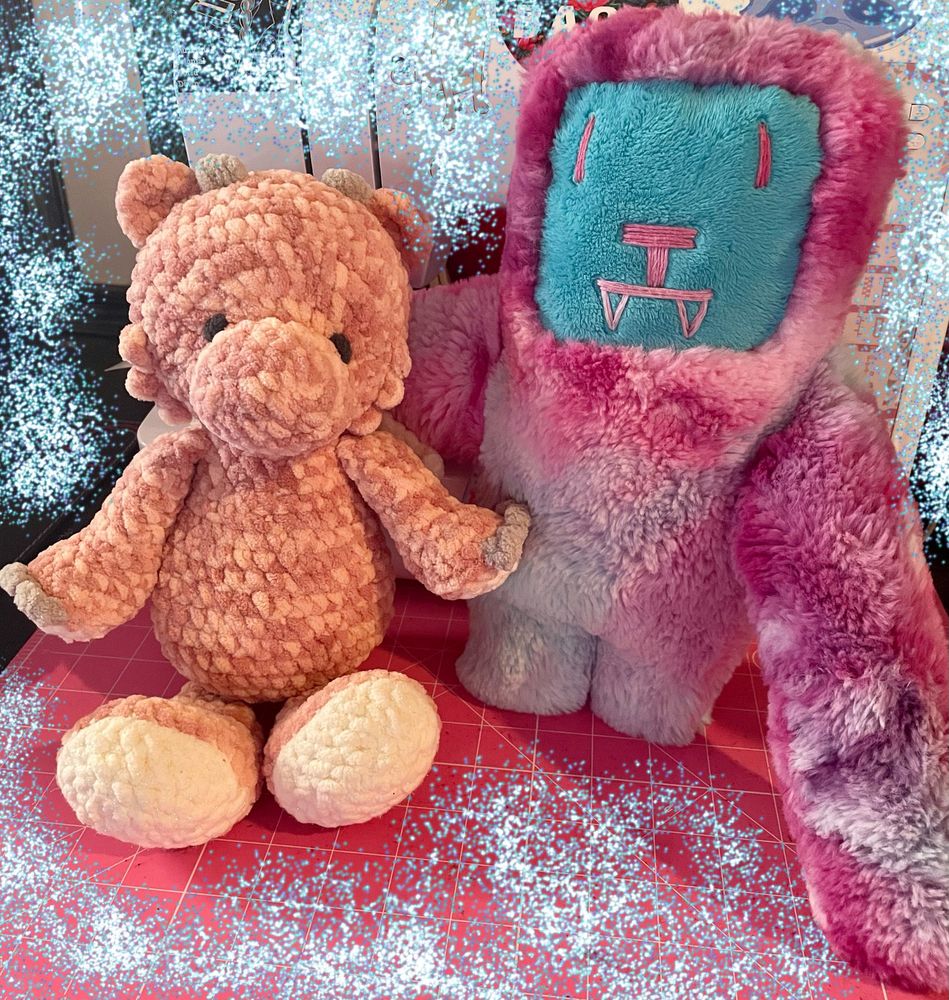 The larger pink dragon next to my own handmade pink and blue plushie. They are about the same size. The background is again obscured by digital blue sparkles. 