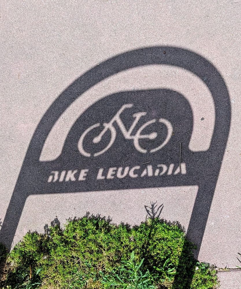A photo of a shadow (on a sidewalk) of a tubular metal bike rack with an inset stenciled cutout image of a bicycle above lettering reading "BIKE LEUCADIA". At the bottom of the photo and shadow is a green patch of weeds.