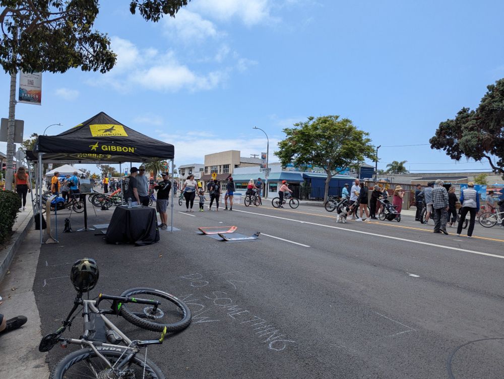 A closed-to-motorized-vehicular-traffic multi-lane coastal highway through downtown Encinitas, CA. A bicycle is on its side at the side of the pavement in the foreground, some street-side trees and tented tables visited by cyclists and pedestrians in the background. The festival is crowned by a light blue sky and a few high, wispy white clouds.