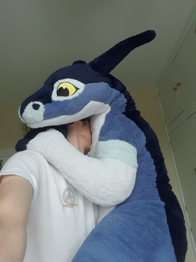 A 2 metre long plush wingless dragon giving his human a big cuddle in celebration of their anniversary together, making sure his human can feel his softness all over. The giant toy dragon is very excited, but his materials mean his face is permanently stuck in a passive neutral expression.