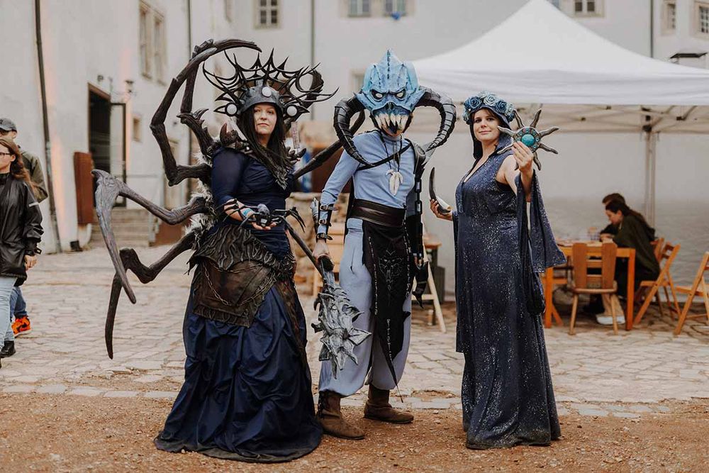 Three cosplayers pose in front of a white tent at ESO Tavern. From left to right: Anonchi as Mephala with spider-like appendages and dark armor; abydoscosplay as Molag Bal in a spiked blue-and-black costume with a mace; and La.Bellaboma as Azura in a starry gown holding a glowing staff. People sit at tables in the background.