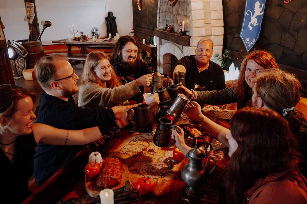 A group of people raising tankards in a toast around a wooden table at the ESO Tavern photo op set. The table is decorated with a map, candles, bread, and tomatoes. Behind them, a stone fireplace and a Daggerfall Covenant banner complete the medieval tavern vibe.