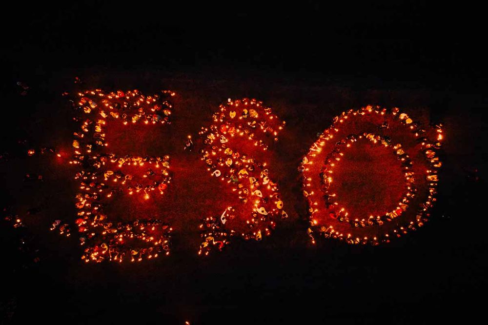 Drone shot at night showing people holding torches that spell out 'ESO' in glowing light against the dark surroundings.