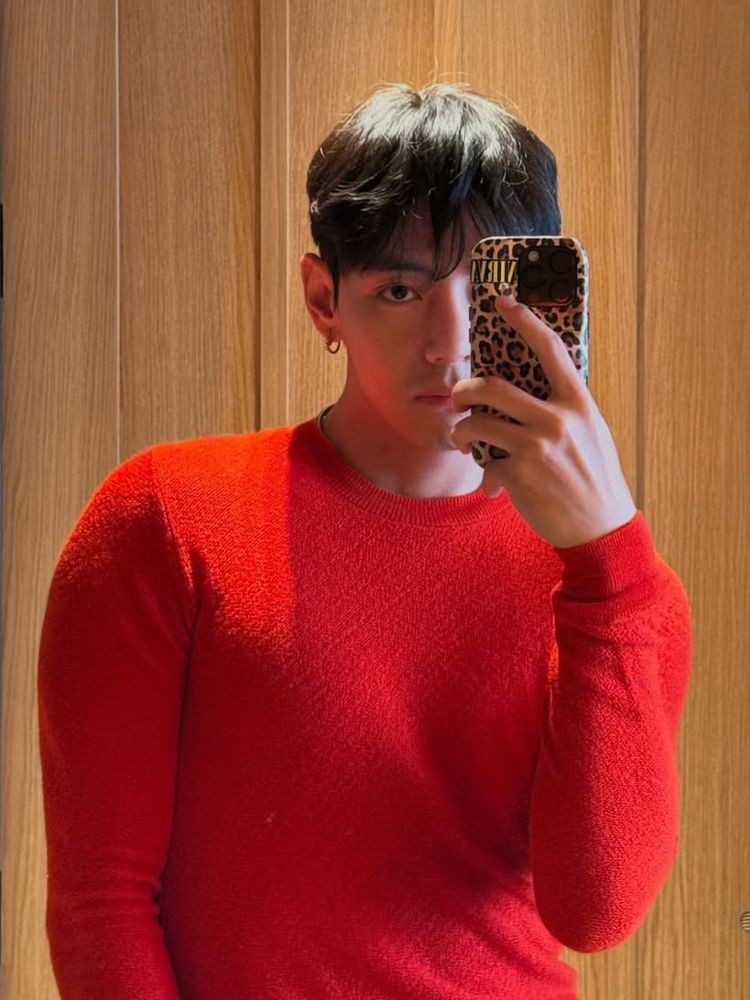 mirror selfie from taehyung’s instagram. he’s wearing a red sweater and has a leopard print phone case, the background is a wood door. 