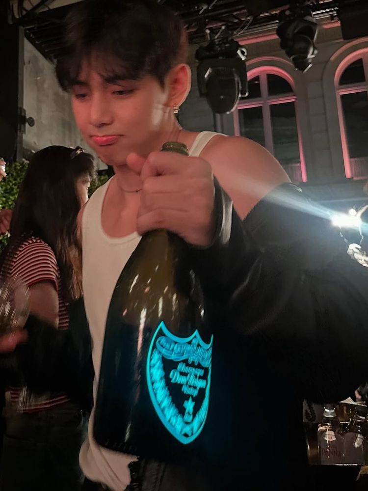 picture from taehyung’s instagram. he is wearing a white tank top and a black jacket, he’s hold in a bottle of liquor and is making a pouty face at a party 