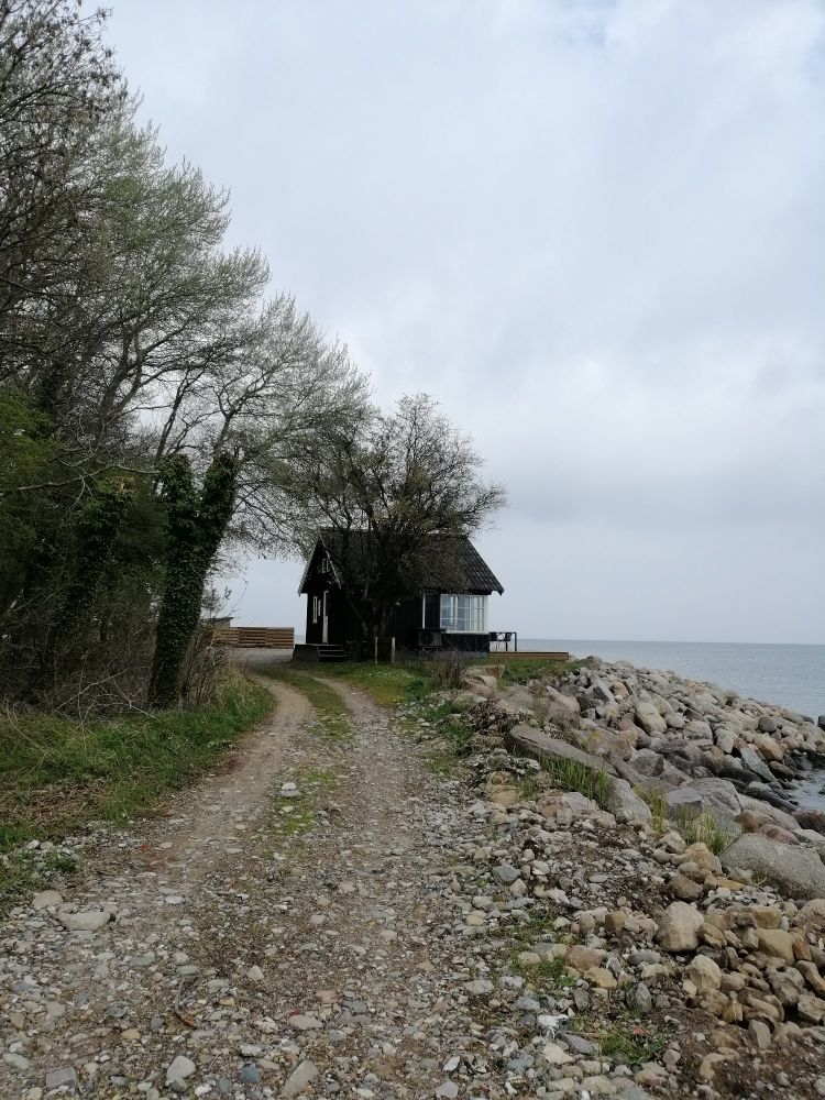 Das Bild zeigt ein kleines, altes Haus am Ende einer einspurigen Schotter Straße. Die Holzterasse ragt über den steinigen Strand hinaus.