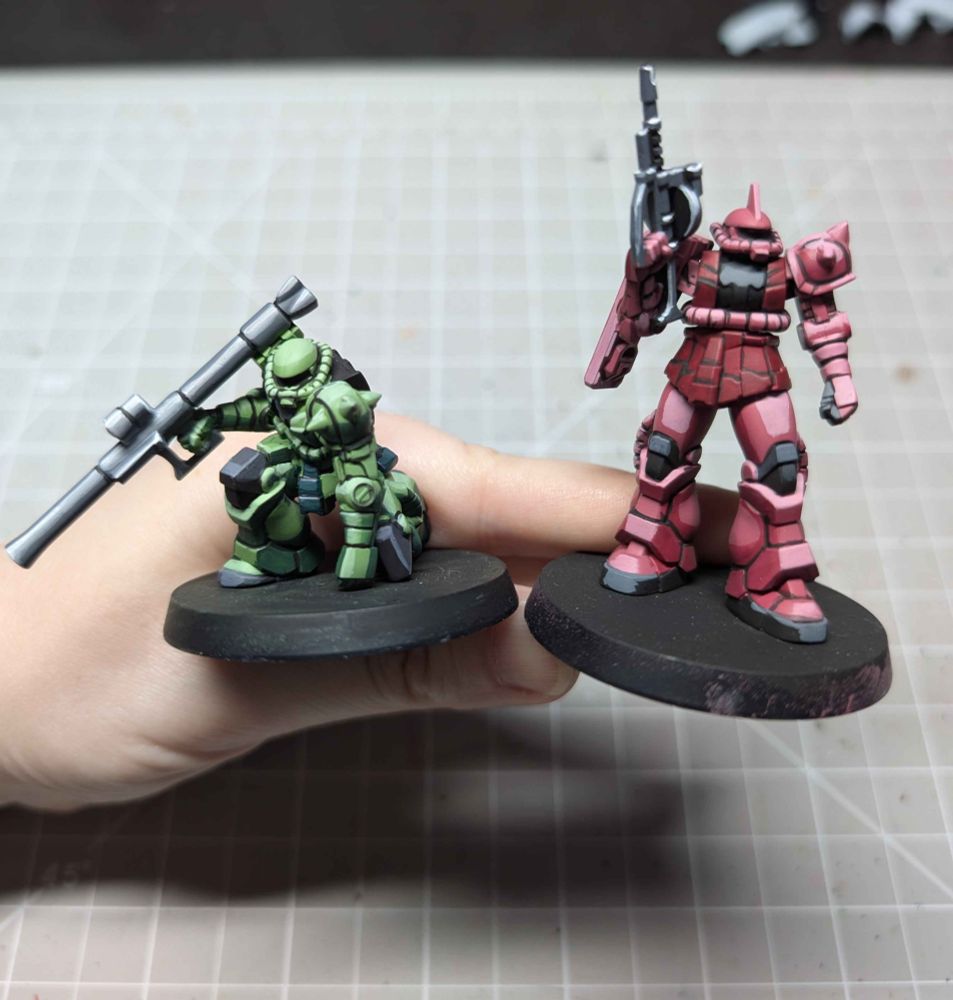 WIP photo of Gundam Assemble miniatures. Left is a Zaku II in green armour painted in a cel-shaded style. Right is Char's Zaku II in pinks and reds also in a cel-shaded style.
