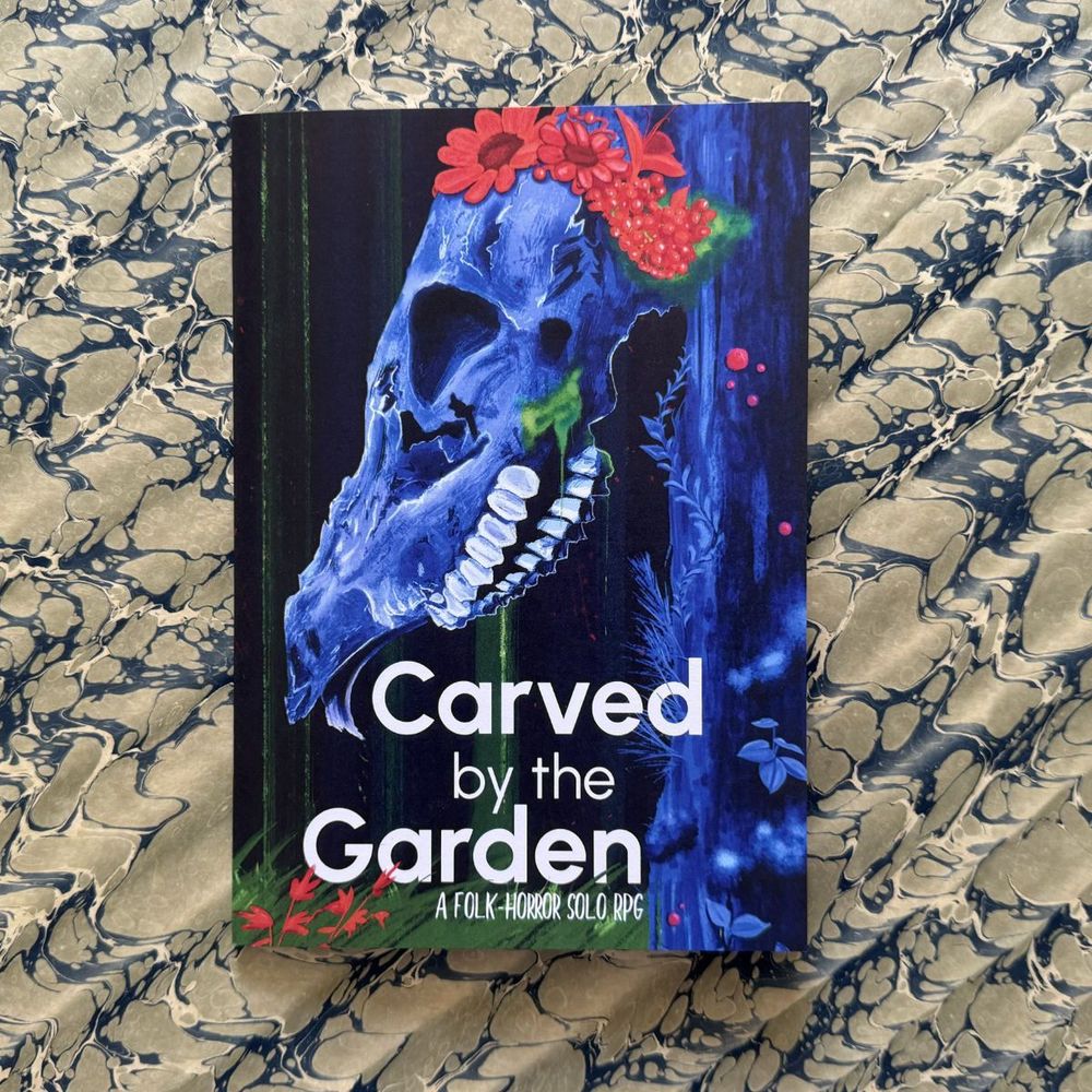 The cover of Carved by the Garden, A folk-horror solo RPG by Cassi Mothwin. A blue-hued deer skull wears red flowers and appears to glow unsettlingly