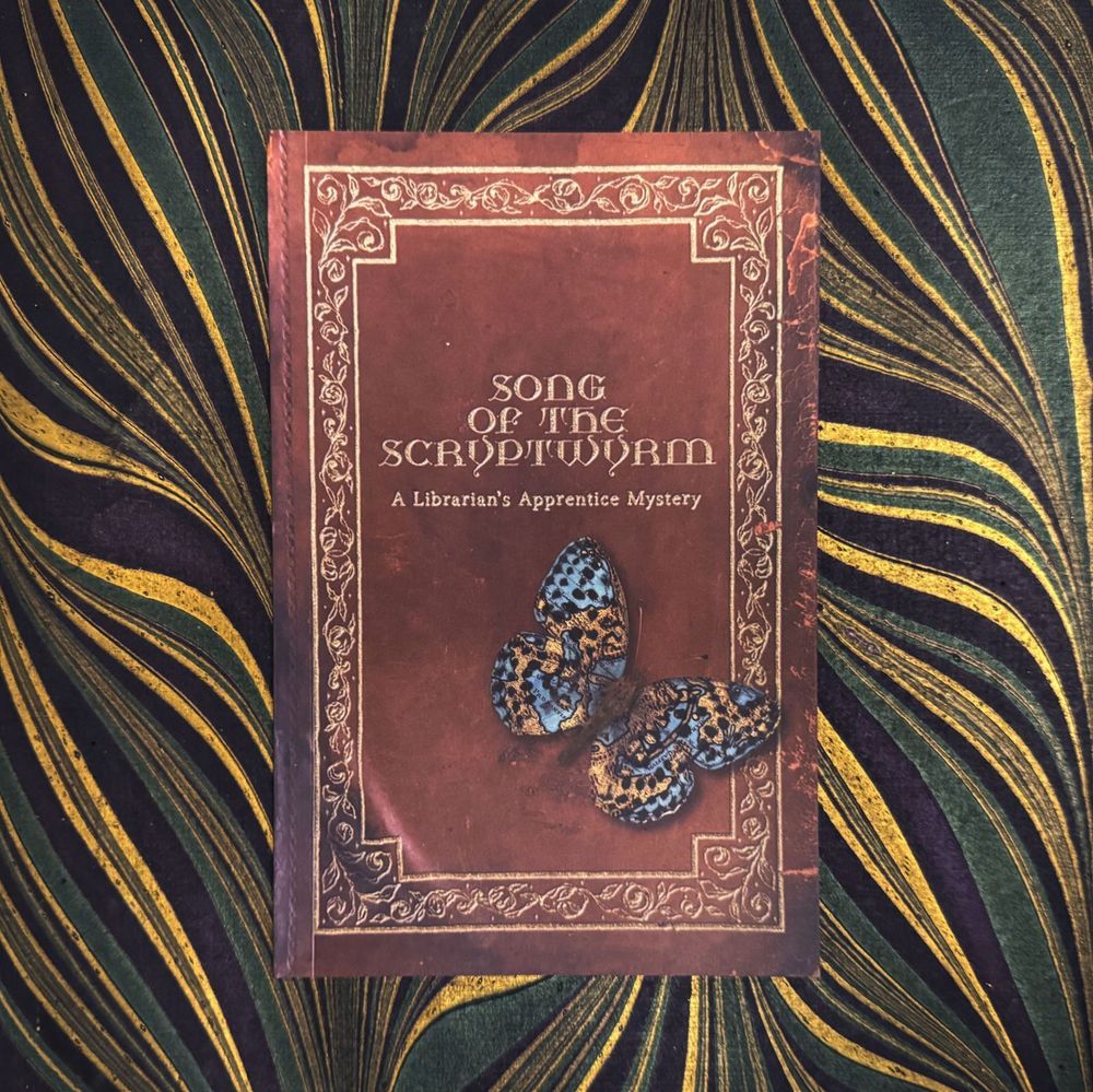 The cover of Song of the Scryptwyrm, A Librarian's Apprentice Mystery. Faux-weathered like a brown leather tome with gold leaf framing embellishment. A photorealistic blue and gold butterfly rests on the bottom right of the cover