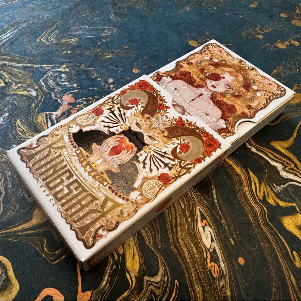 The box for the "Light" version of King Star Silence V3 Twins playing cards. White background with gold foil embellishment. The box is arranged to reveal an outer sleeve with laser-cut sections meant to display the inner sleeve (which holds the cards). A gold-haired woman is visible on the inner box. Together, the outer sleeve adds her hands holding a gemstone heart and gives her a ram's horn masquerade mask