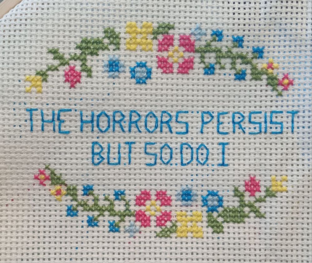 Flowery cross-stitch in green, yellow, pink and blue surrounds the juxtaposed stitched message, "The horrors persist but so do I". Credit: secretsfromwholecloth